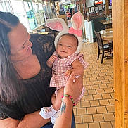 Aurora is registered to the contest to win money with this photo: baby, child, woman, bunny_ears, pink_clothing, restaurant, indoor, tiled_floor, smile, holding, person, socks, tattoo, bracelet, table, chair, window, natural_light, happy, cute