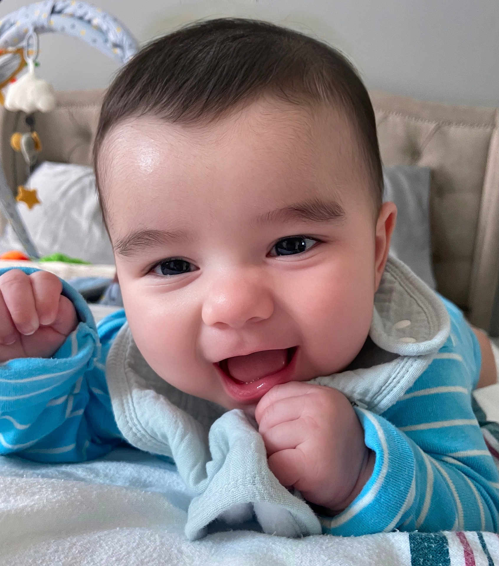 Aaron is registered to the contest to win money with this photo: baby, smiling, close_up, face, blue_clothing, bib, hand, indoor, toy, bed, blanket, happy, cute, infant, person, child, skin, head, expression, toddler