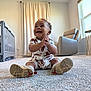 infant, baby, smiling, sitting, clapping_hands, onesie, patterned_clothing, sneakers, shoe_soles, carpet, nursery, crib, armchair, curtains, window, soft_light, indoor, happy, portrait, playful
