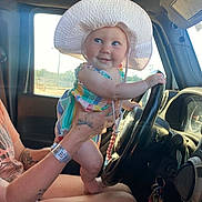 Hazel joined the competition — help win amazing prizes! baby, child, hat, sunhat, steering_wheel, car_interior, tattoo, adult_hand, hospital_wristband, window, sunlight, smile, colorful_clothing, person, seat, dashboard, keys, playful, cute, infant