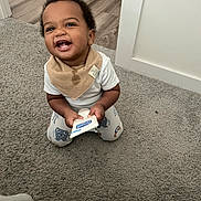 Reece joined the competition — help win amazing prizes! baby, child, infant, smiling, laughing, sitting, carpet, indoors, home, bib, white_shirt, patterned_pants, toy, package, doorway, wood_floor, portrait, happy, curly_hair, teeth