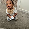 baby, child, infant, smiling, laughing, sitting, carpet, indoors, home, bib, white_shirt, patterned_pants, toy, package, doorway, wood_floor, portrait, happy, curly_hair, teeth