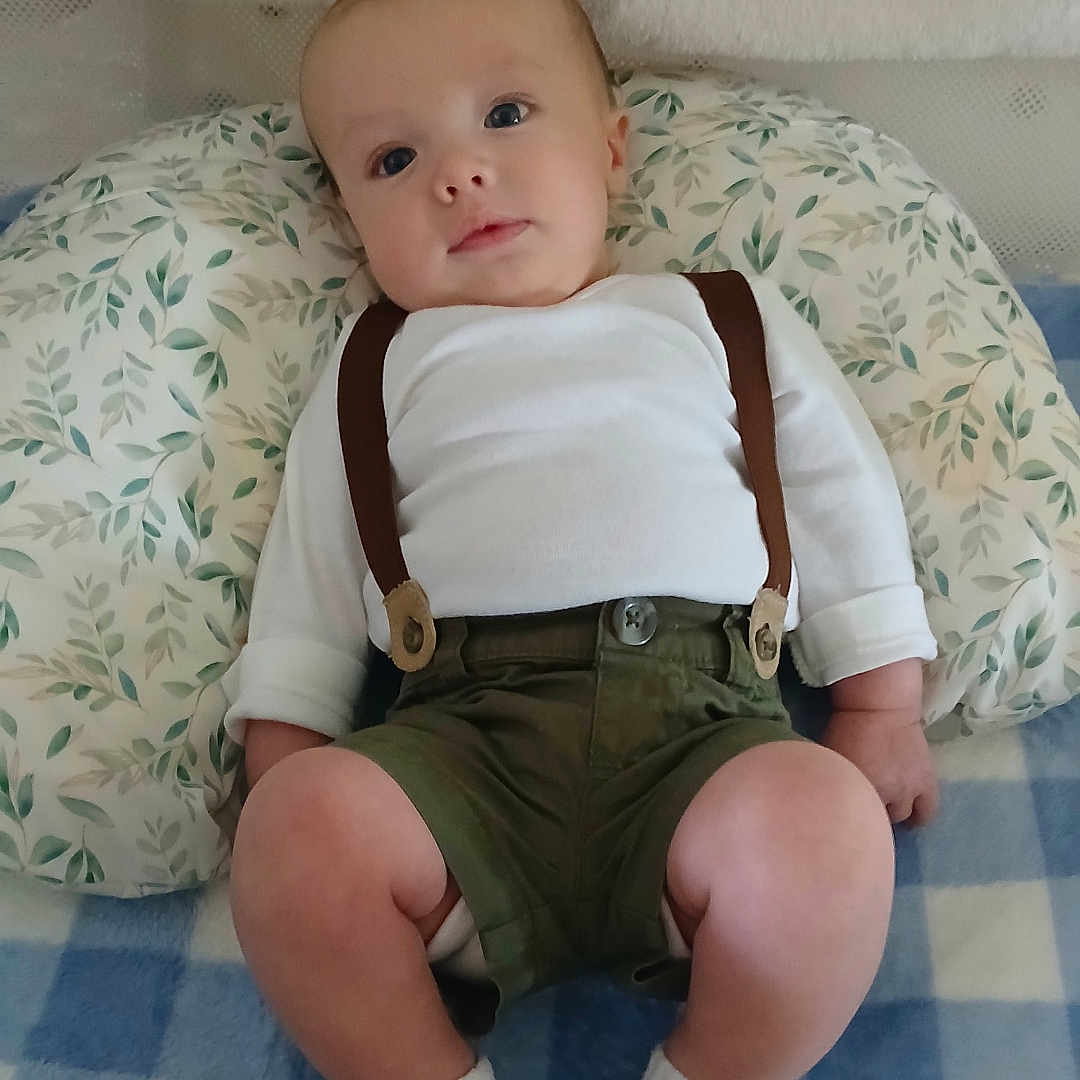 Kasyn is registered to the contest to win money with this photo: baby, infant, child, portrait, lying_down, sitting, suspenders, white_shirt, green_shorts, socks, pillow, blanket, crib, nursery, indoor, face, eyes, curious, patterned_fabric, photography