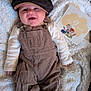 baby, smiling, corduroy_overalls, hat, blanket, milestone_card, footwear, infant, clothing, happy, indoor, portrait, cute, child, baby_shoes, face, person, resting, young_child, soft_texture