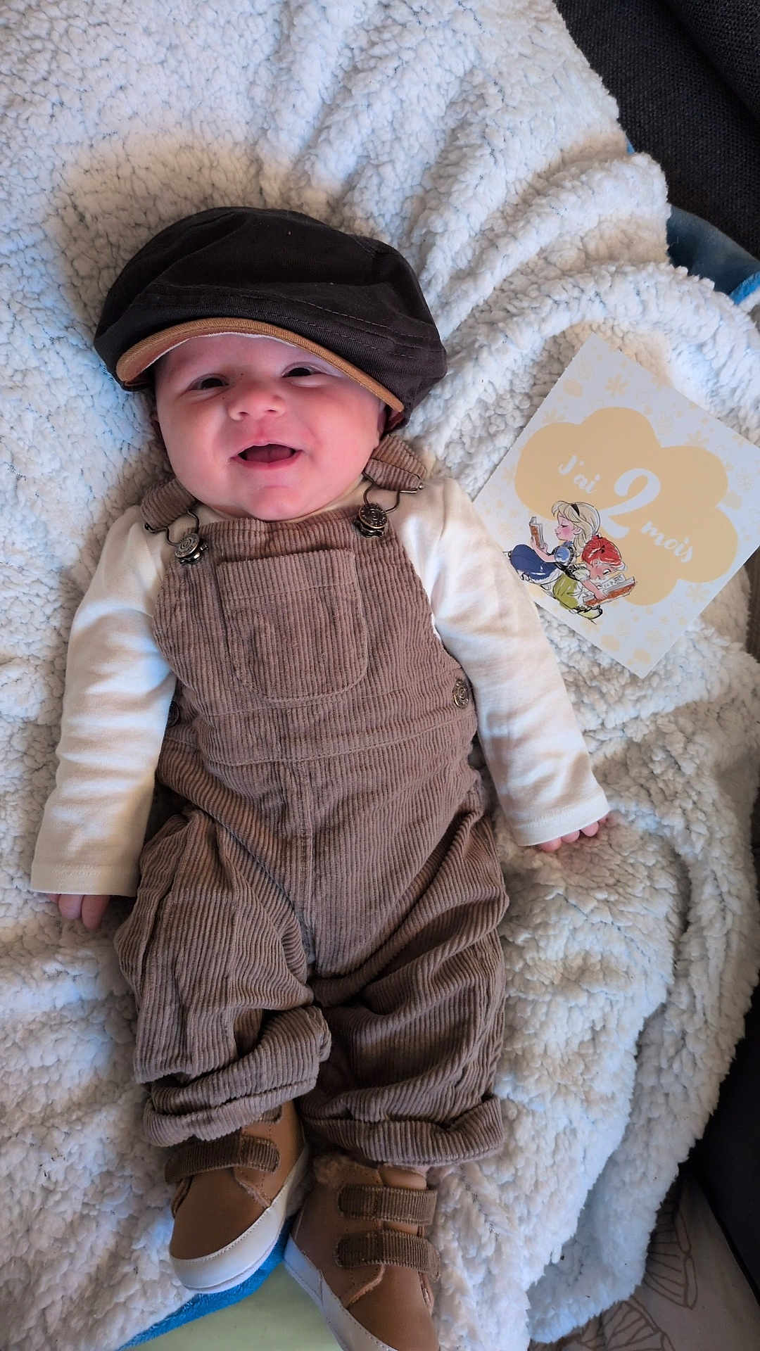 Abygaël participe au concours pour gagner de l'argent avec cette photo : baby, smiling, corduroy_overalls, hat, blanket, milestone_card, footwear, infant, clothing, happy, indoor, portrait, cute, child, baby_shoes, face, person, resting, young_child, soft_texture