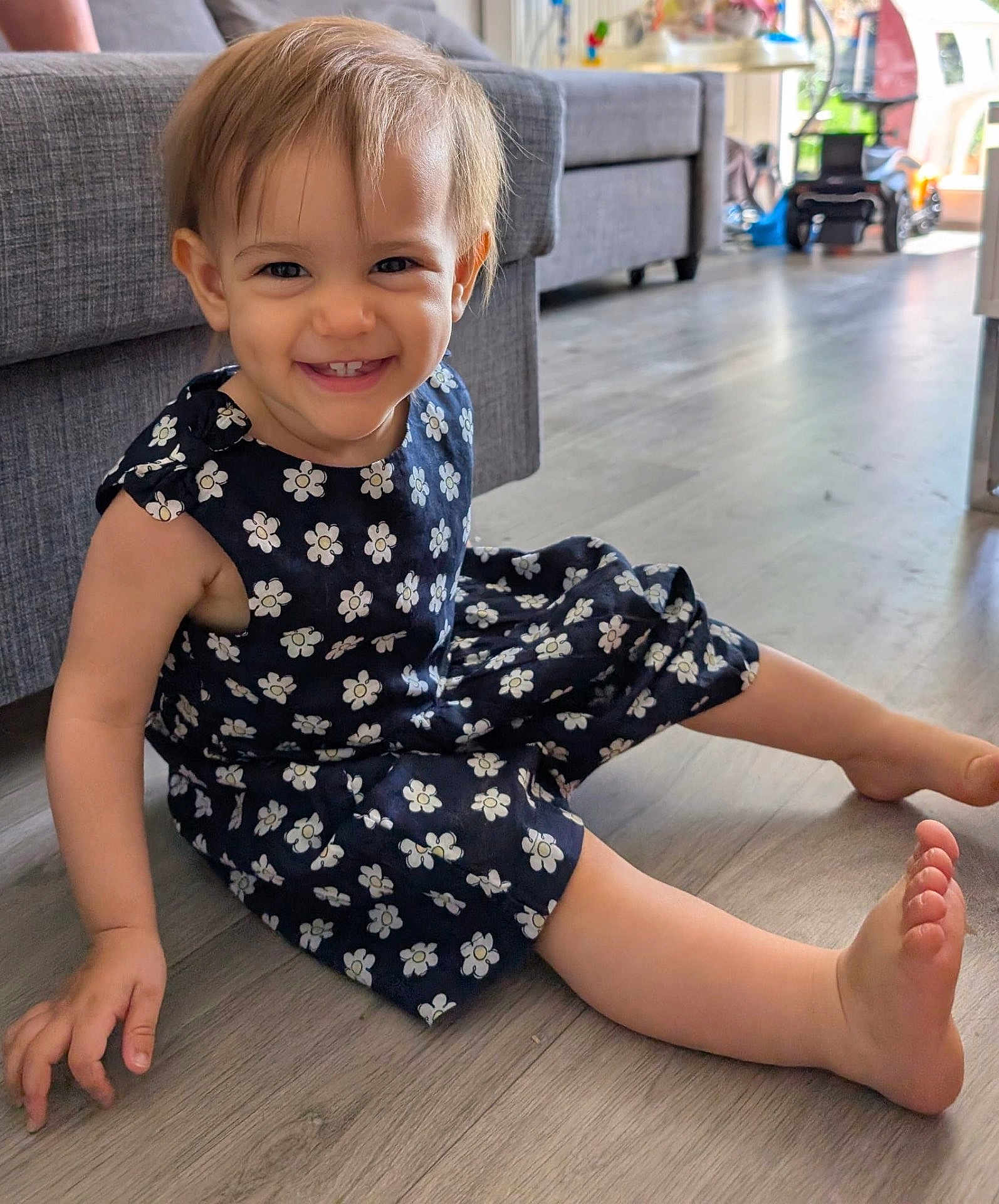 Soleya a rejoint le concours — aidez-le/la à gagner de superbes lots ! toddler, child, smiling, sitting, dress, flower_pattern, barefoot, floor, wooden_floor, couch, indoor, home, happy, person, young_child, furniture, living_room, cute, portrait, casual