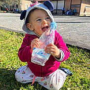 Soleya a rejoint le concours — aidez-le/la à gagner de superbes lots ! child, toddler, grass, outdoor, water_bottle, hood, jacket, smiling, happy, cute, sunny_day, building, people_in_background, casual_clothing, playful, nature, daylight, kneeling, pink, ear_hood
