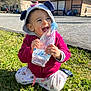 child, toddler, grass, outdoor, water_bottle, hood, jacket, smiling, happy, cute, sunny_day, building, people_in_background, casual_clothing, playful, nature, daylight, kneeling, pink, ear_hood