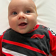 Maddox is registered to the contest to win money with this photo: baby, infant, smiling, face, eyes, clothing, red, black, white, racing_theme, formula_1, chequered_flag, shirt, cute, happy, portrait, closeup, lying_down, soft_background, child