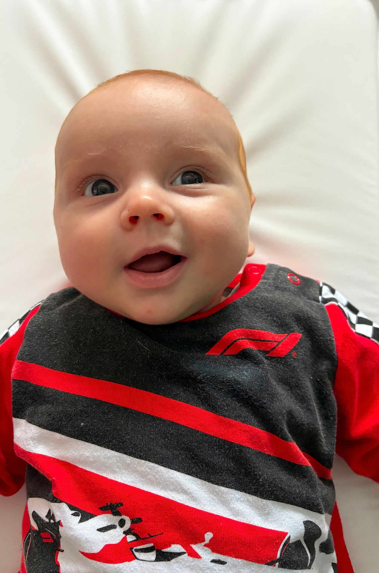 Maddox is registered to the contest to win money with this photo: baby, infant, smiling, face, eyes, clothing, red, black, white, racing_theme, formula_1, chequered_flag, shirt, cute, happy, portrait, closeup, lying_down, soft_background, child