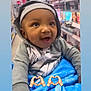 baby, child, hoodie, blanket, smiling, indoor, shopping_aisle, sitting, person, face, cute, happy, clothing, earrings, headwear, blue, gray, store, retail, emojis