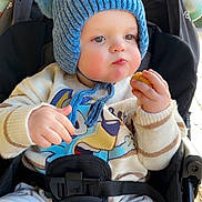 Cody is registered to the contest to win money with this photo: baby, blue_hat, cartoon_clothing, child, cozy, cute, daylight, expression, face, food, hands, outdoor, plush, pom_poms, seatbelt, sitting, snack, stroller, sweater, toddler