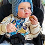 baby, blue_hat, cartoon_clothing, child, cozy, cute, daylight, expression, face, food, hands, outdoor, plush, pom_poms, seatbelt, sitting, snack, stroller, sweater, toddler