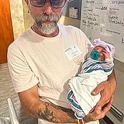 Karolina is registered to the contest to win money with this photo: man, baby, newborn, hospital, pacifier, beard, glasses, hat, tattoo, white_shirt, hospital_room, blanket, arm, holding, infant, portrait, headwear, indoor, person, medical