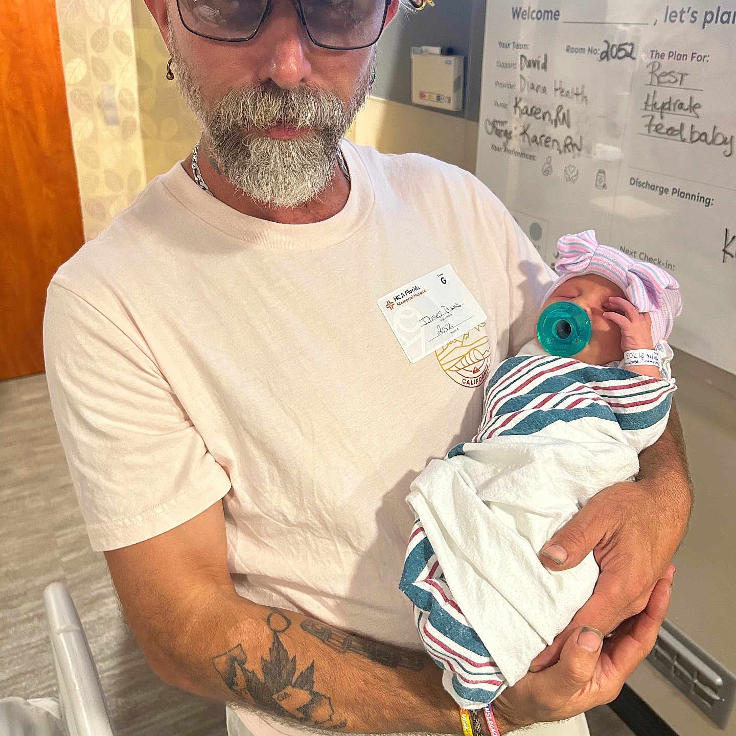 Karolina is registered to the contest to win money with this photo: man, baby, newborn, hospital, pacifier, beard, glasses, hat, tattoo, white_shirt, hospital_room, blanket, arm, holding, infant, portrait, headwear, indoor, person, medical