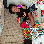Jaeluv is registered to the contest to win money with this photo: toddler, child, curly_hair, playpen, toys, colorful, smiling, happy, indoor, plastic_toys, stuffed_animal, pop_it_toy, book, foot, black_clothing, play_area, baby, fun, cute, early_childhood