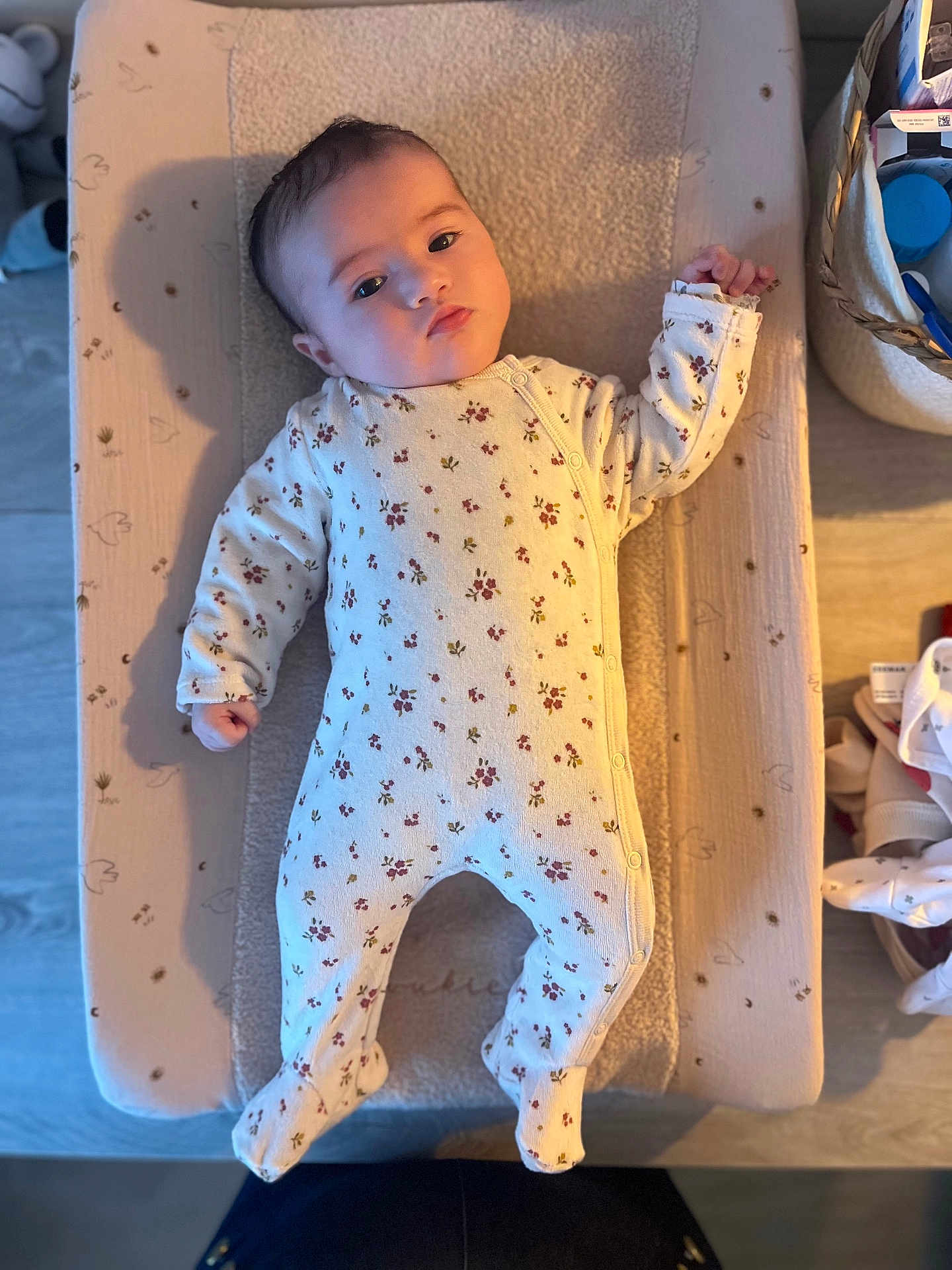 Romane participe au concours pour gagner de l'argent avec cette photo : baby, infant, onesie, floral_pattern, changing_pad, shadow, face, hand, footed_pajamas, indoor, wooden_floor, basket, household_items, soft_texture, calm, lying_down, person, cute, child, newborn