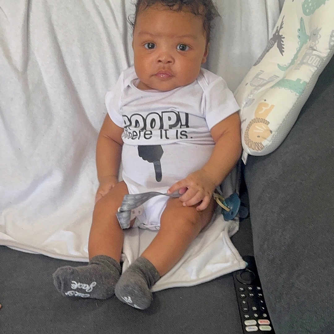 Grayson is registered to the contest to win money with this photo: baby, child, infant, couch, blanket, onesie, socks, remote_control, indoor, furniture, person, seated, curious_expression, hair, skin, face, hand, foot, white, gray