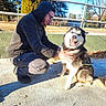 canine, casual_clothing, crouching, daylight, dog, fence, friendship, glasses, happy, hoodie, husky, man, nature, outdoor, park, pavement, pet, smiling, sunlight, trees