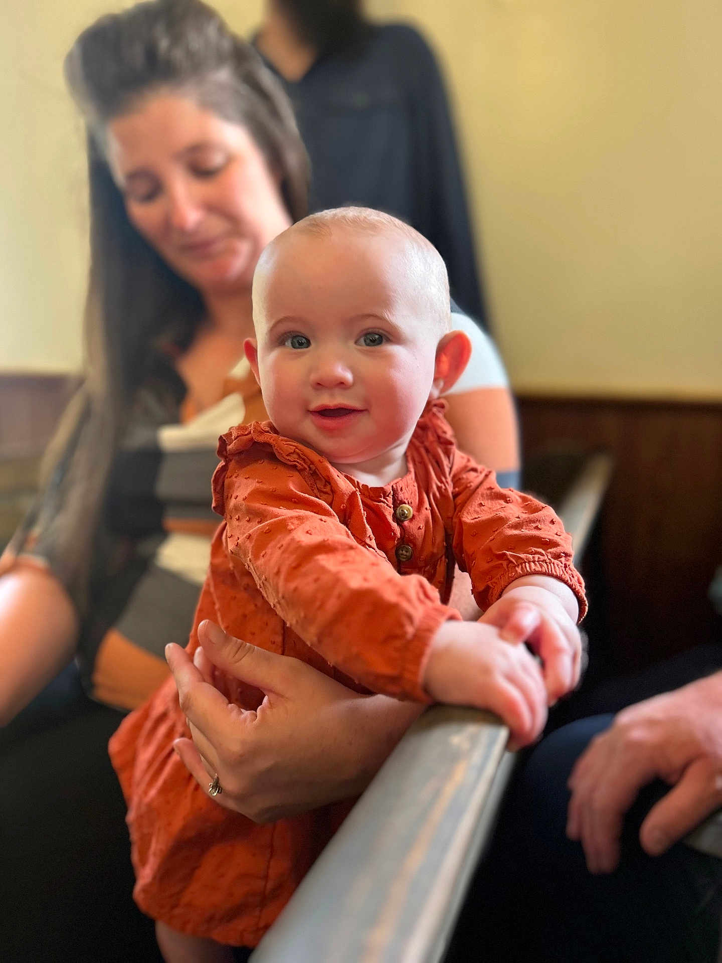 Avery joined the competition — help win amazing prizes! baby, child, woman, adult, orange_dress, holding, smile, indoor, person, face, hand, curious, seated, blurred_background, wood_paneling, long_hair, casual_clothing, skin, portrait, family