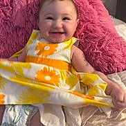Emily is registered to the contest to win money with this photo: baby, child, smiling, portrait, indoor, sitting, pink_blanket, fluffy_cushion, floral_dress, yellow, orange, white, diaper, bed, home, happy, face, arms, legs, cozy