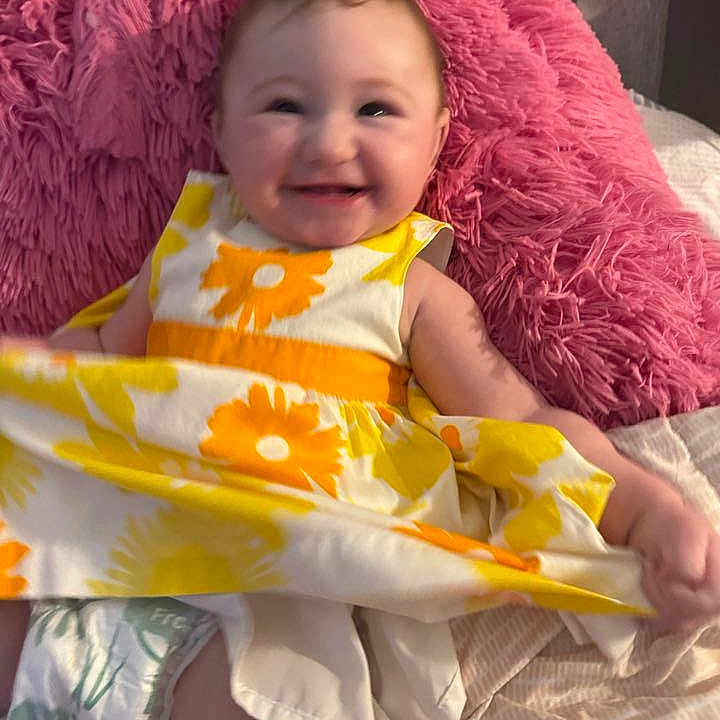 Emily is registered to the contest to win money with this photo: baby, child, smiling, portrait, indoor, sitting, pink_blanket, fluffy_cushion, floral_dress, yellow, orange, white, diaper, bed, home, happy, face, arms, legs, cozy
