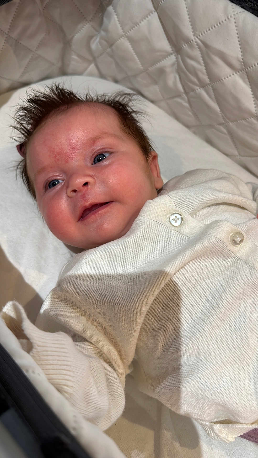 Evelyn joined the competition — help win amazing prizes! baby, infant, face, expression, smile, hair, blue_eyes, white_clothing, quilted_background, skin, newborn, cute, lying_down, soft_texture, child, happy, portrait, indoors, buttoned_clothes, baby_garment