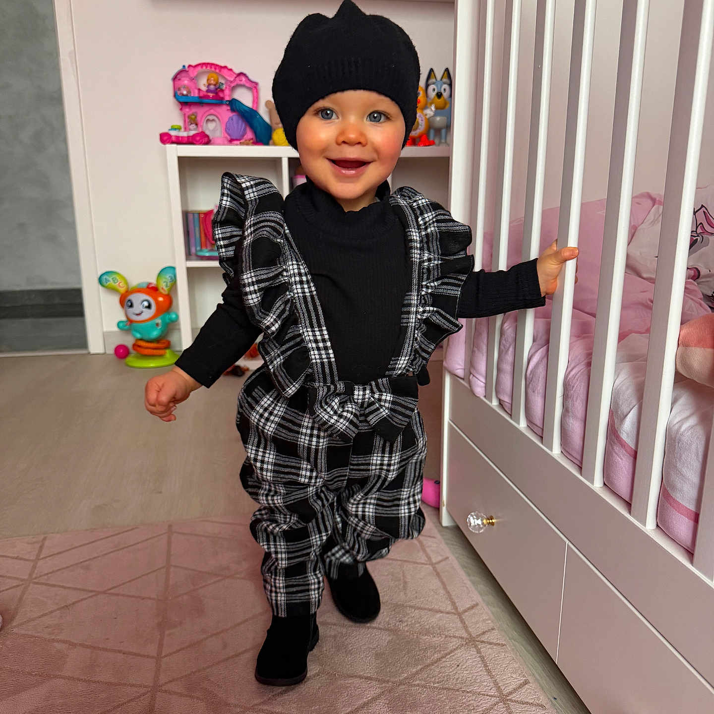 Molly participe au concours pour gagner de l'argent avec cette photo : area_rug, beanie, black_clothing, child, crib, cute, doll, flooring, hat, indoor, nursery, pink_bedding, plaid_overalls, portrait, ruffles, shelf, smile, toddler, toy, walking