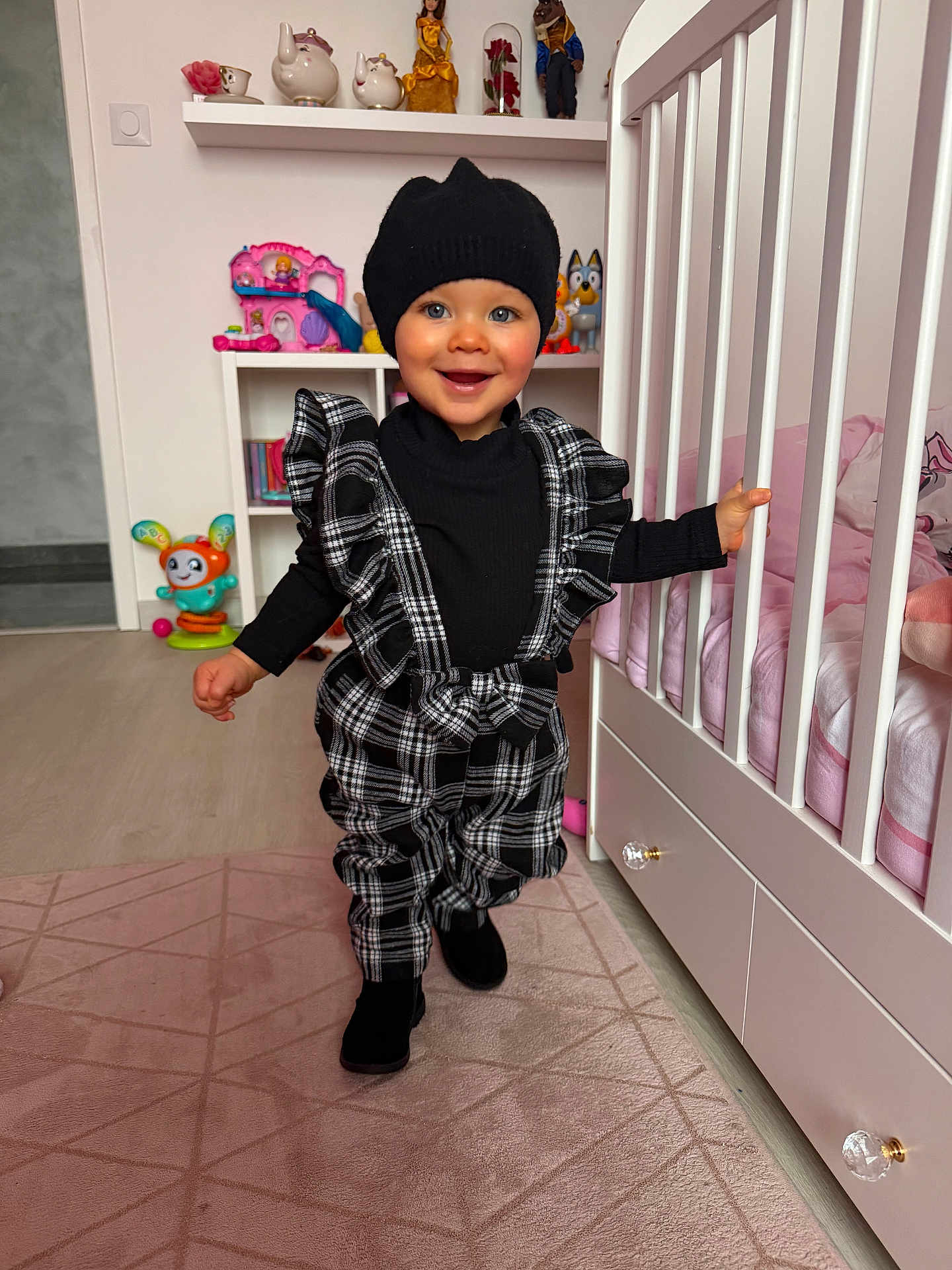 Molly participe au concours pour gagner de l'argent avec cette photo : child, toddler, smile, beanie, hat, plaid_overalls, ruffles, black_clothing, crib, nursery, toy, doll, shelf, pink_bedding, area_rug, flooring, portrait, walking, cute, indoor