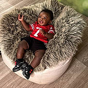 Maaz is registered to the contest to win money with this photo: baby, child, smiling, red_jersey, black_shorts, bean_bag, fluffy, furniture, flooring, happy, indoor, footwear, sneakers, person, cute, infant, relaxed, portrait, young_child, cozy