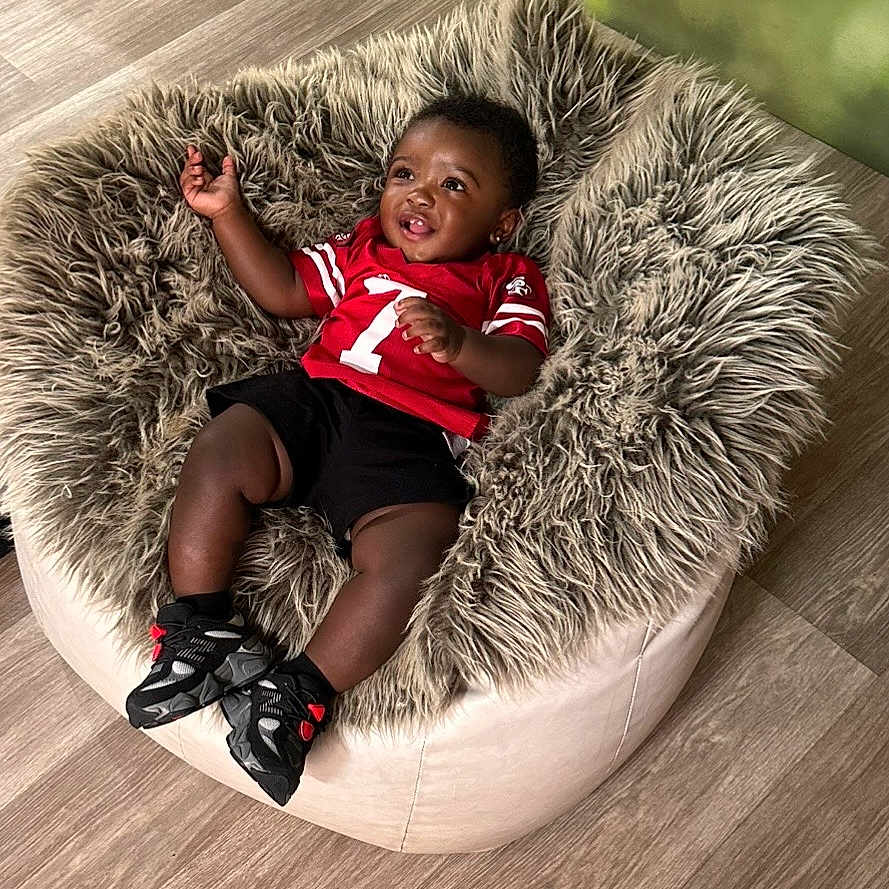 Maaz is registered to the contest to win money with this photo: baby, child, smiling, red_jersey, black_shorts, bean_bag, fluffy, furniture, flooring, happy, indoor, footwear, sneakers, person, cute, infant, relaxed, portrait, young_child, cozy