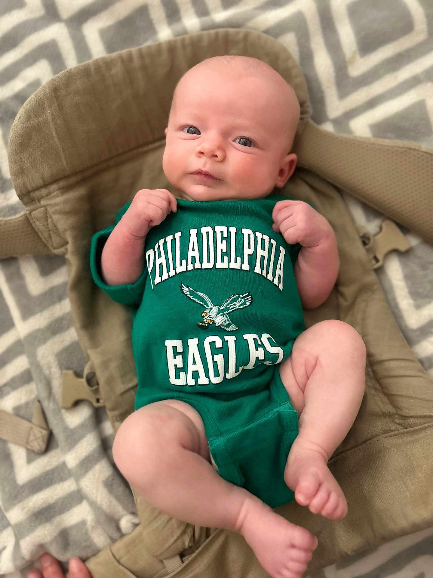 Sophia is registered to the contest to win money with this photo: baby, infant, onesie, green_clothing, philadelphia_eagles, baby_carrier, blanket, patterned_blanket, foot, hand, face, head, skin, person, lying_down, indoor, closeup, cute, newborn, comfort