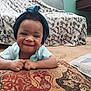 baby, smiling, head_wrap, cushion, blanket, couch, carpet, indoor, child, happy, person, patterned, cozy, living_room, portrait, infant, face, hands, clothing, home