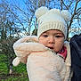baby, child, winter_clothing, hat, pom_poms, jacket, outdoor, trees, sky, person, face, cute, cold_weather, nature, greenery, portrait, expression, infant, holding, warm_clothes