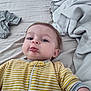 baby, child, pout, yellow_stripes, onesie, bed, blanket, clothing, face, person, infant, rumpled_bed, lying_down, cute, expression, indoors, toddler, sleepwear, soft_lighting, casual