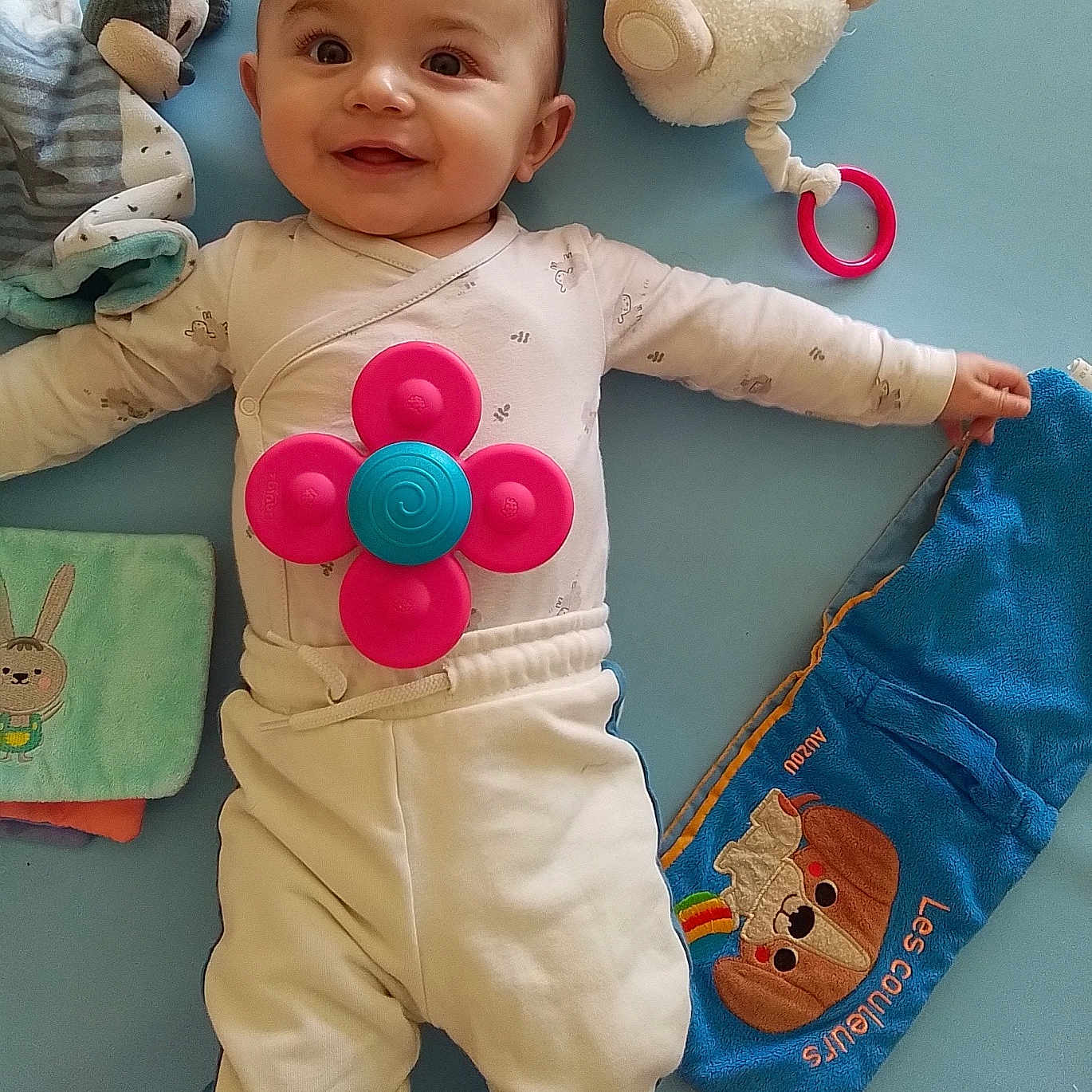 Aylan participe au concours pour gagner de l'argent avec cette photo : baby, smiling, toy, blue_background, clothing, infant, lying_down, cute, happy, playful, child, soft_toy, colorful, fabric, casual_wear, indoors, person, small_hand, bright_eyes, cheerful