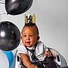 Marley is registered to the contest to win money with this photo: baby, child, high_chair, crown, balloon, black_balloon, white_balloon, blue_balloon, tie, shirt, smiling, celebration, portrait, indoor, happy, person, clothing, sitting, cute, half_birthday