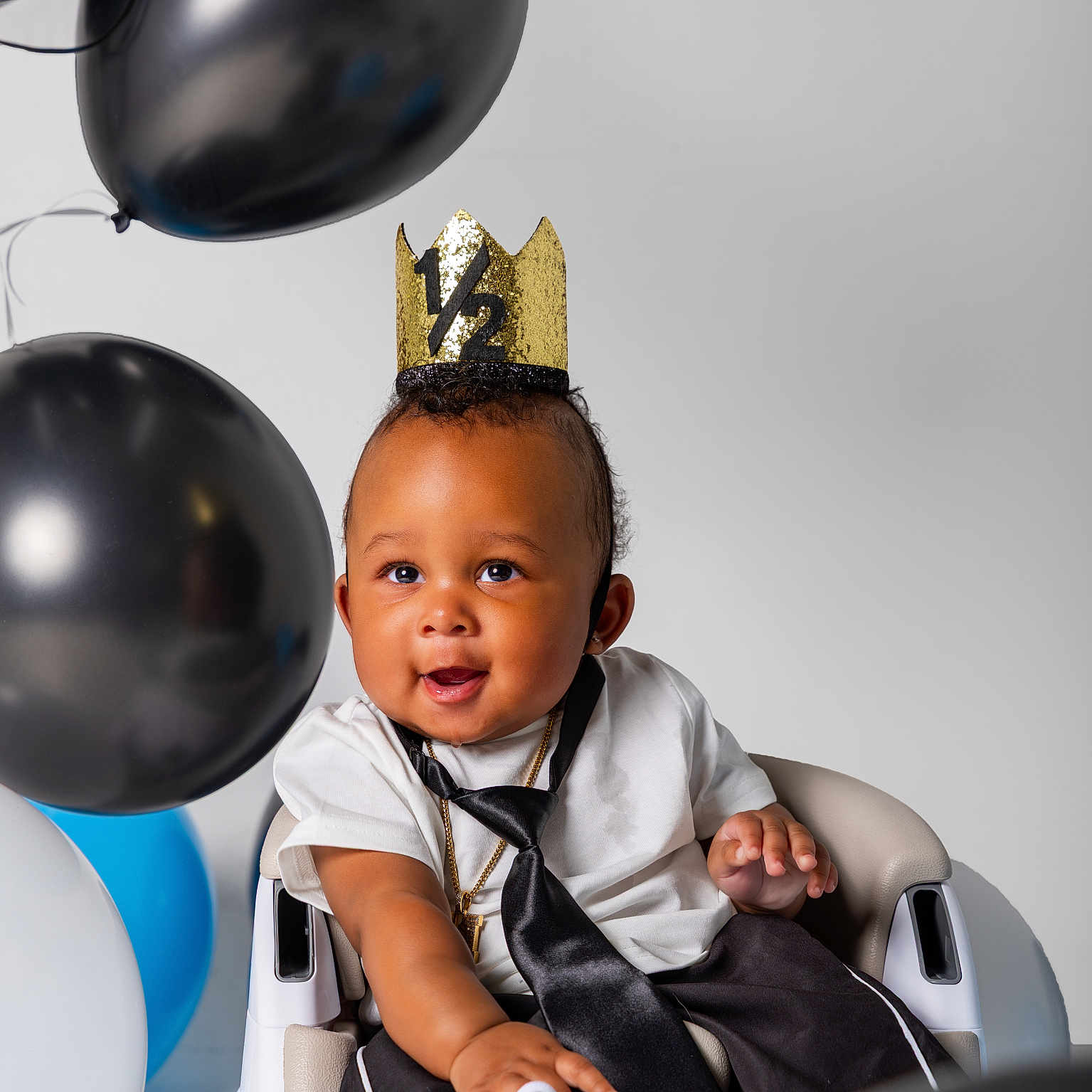 Marley is registered to the contest to win money with this photo: baby, child, high_chair, crown, balloon, black_balloon, white_balloon, blue_balloon, tie, shirt, smiling, celebration, portrait, indoor, happy, person, clothing, sitting, cute, half_birthday