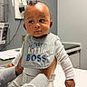 baby, infant, child, bib, mister_little_boss, tattooed_hands, clothing, white_long_sleeve, gray_pants, indoor, medical_office, table, curious_expression, person_holding_baby, face, skin, head, portrait, sitting, furniture