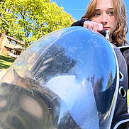 Bowie is registered to the contest to win money with this photo: woman, person, face, portrait, pet_carrier, animal_carrier, outdoor, suburban, house, tree, sunlight, blue_sky, road, sidewalk, grass, backpack, black_clothing, brown_hair, looking_at_camera, pet