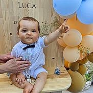 Luca a rejoint le concours — aidez-le/la à gagner de superbes lots ! baby, child, bow_tie, white_shirt, shorts, balloons, wooden_bench, hand, flowers, celebration, pointing, face, legs, feet, outdoor, party, decorations, pacifier_chain, smile, person