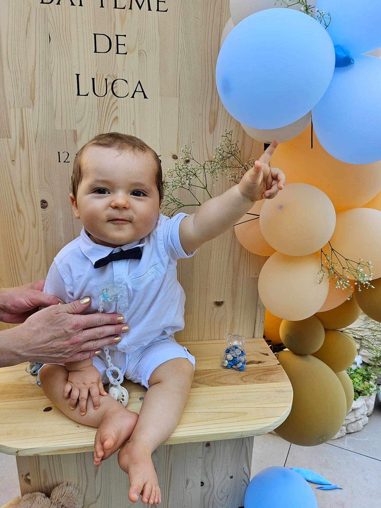 Luca a rejoint le concours — aidez-le/la à gagner de superbes lots ! baby, child, bow_tie, white_shirt, shorts, balloons, wooden_bench, hand, flowers, celebration, pointing, face, legs, feet, outdoor, party, decorations, pacifier_chain, smile, person