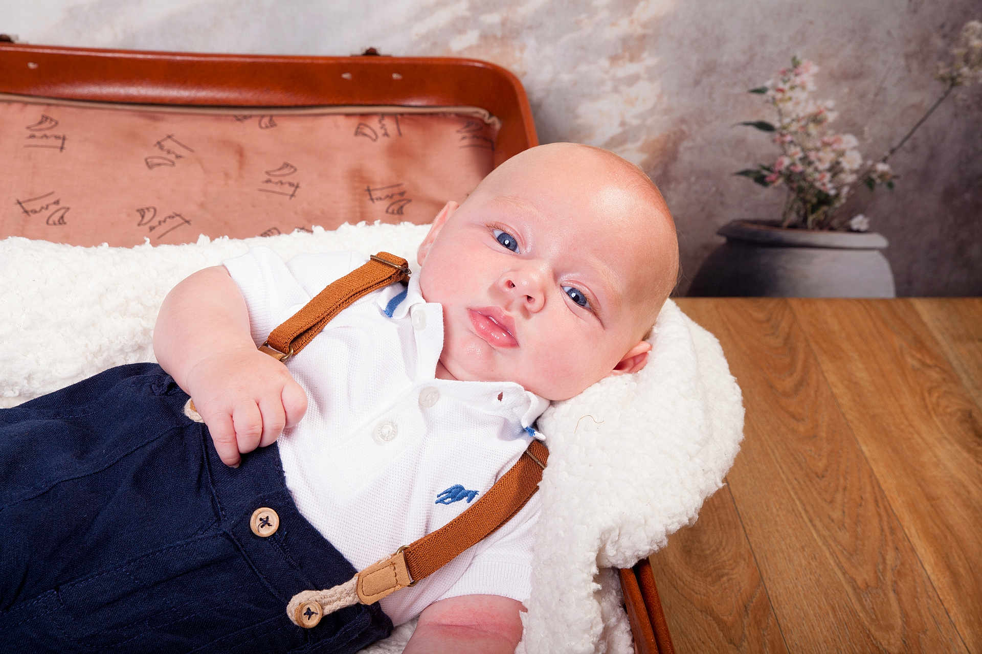 Thibault participe au concours pour gagner de l'argent avec cette photo : baby, infant, blue_eyes, white_shirt, suspenders, blanket, vintage_suitcase, wooden_table, flower_vase, indoor, portrait, cute, chubby_cheeks, lying_down, soft_texture, casual_clothing, skin, head, hand, relaxation