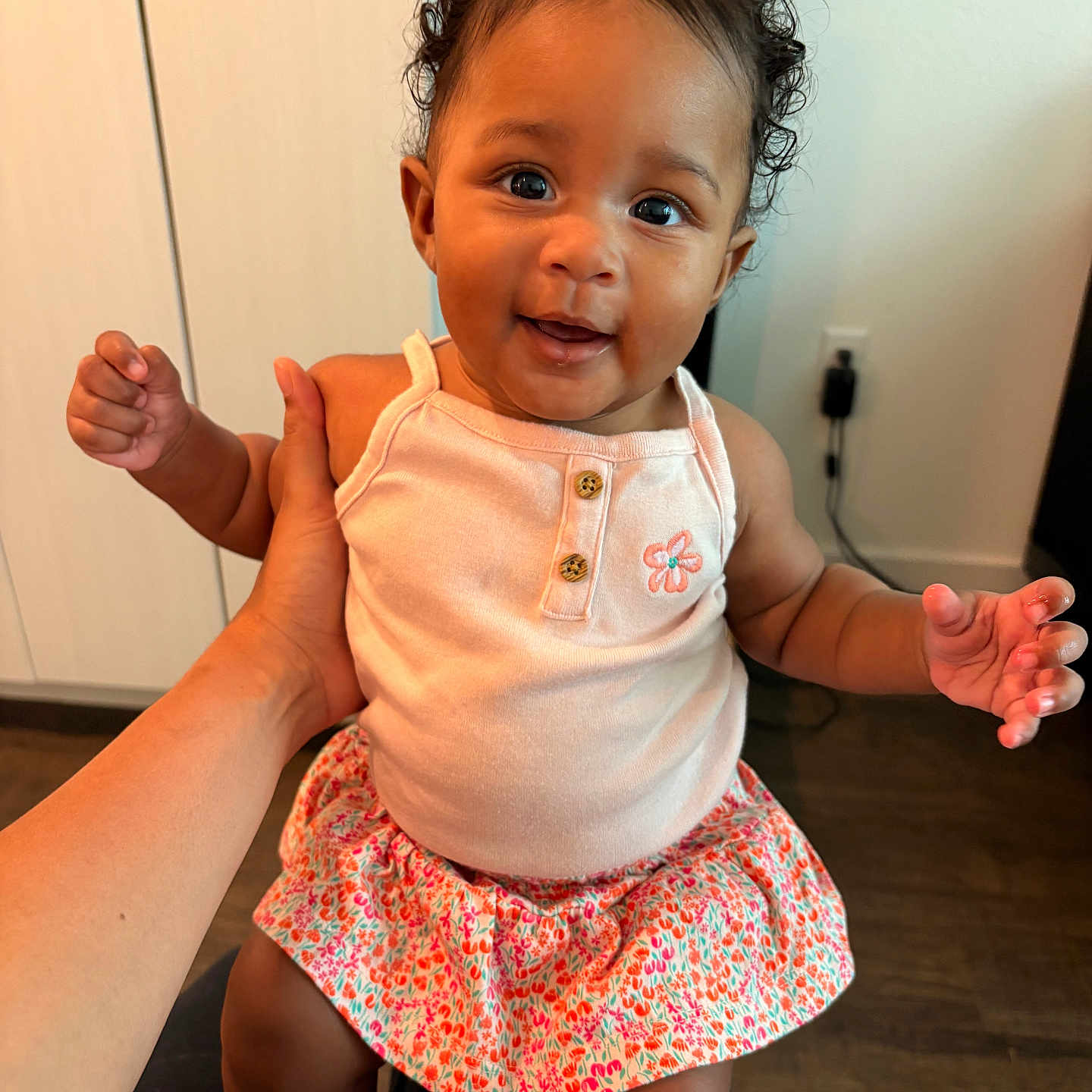 Azelia is registered to the contest to win money with this photo: baby, child, smiling, curly_hair, pink_top, floral_skirt, indoor, hand, person, cute, happy, portrait, seated, skin, eyes, clothing, furniture, floor, cabinet, light