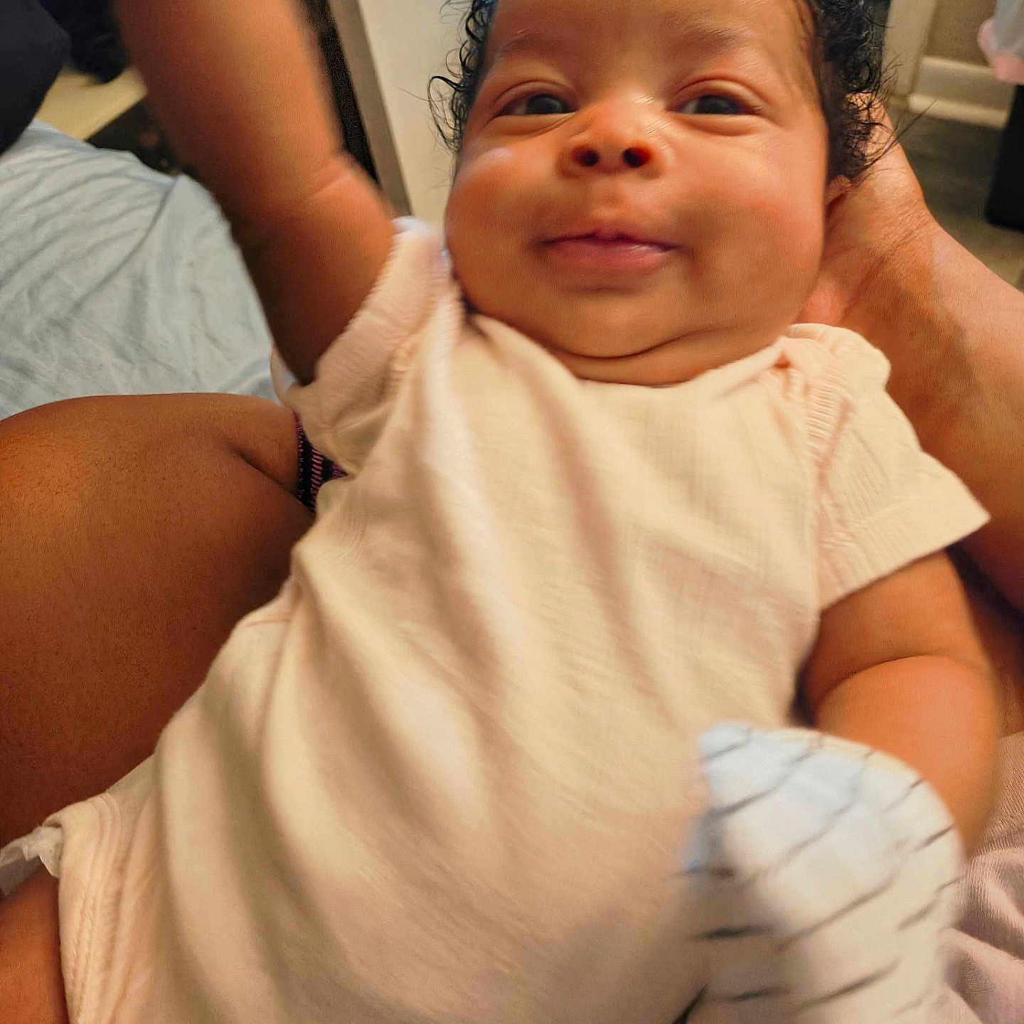 Saphira is registered to the contest to win money with this photo: baby, infant, child, curly_hair, onesie, mittens, smiling, happy, arm, hand, person, indoor, bedroom, adult_hand, skin, couch, fabric, closeup, portrait, playful