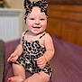 baby, infant, smiling, sitting, leopard_print, headband, bow, clothing, indoor, portrait, child, happy, furniture, purple_bedspread, skin, cute, young_child, person, face, feet