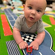 Daniel is registered to the contest to win money with this photo: baby, infant, child, toy_car, play_mat, racetrack, carpet, crawling, hands, closeup, portrait, big_eyes, cute, indoor, soft_lighting, bokeh, foreground, colorful, checkerboard, onesie