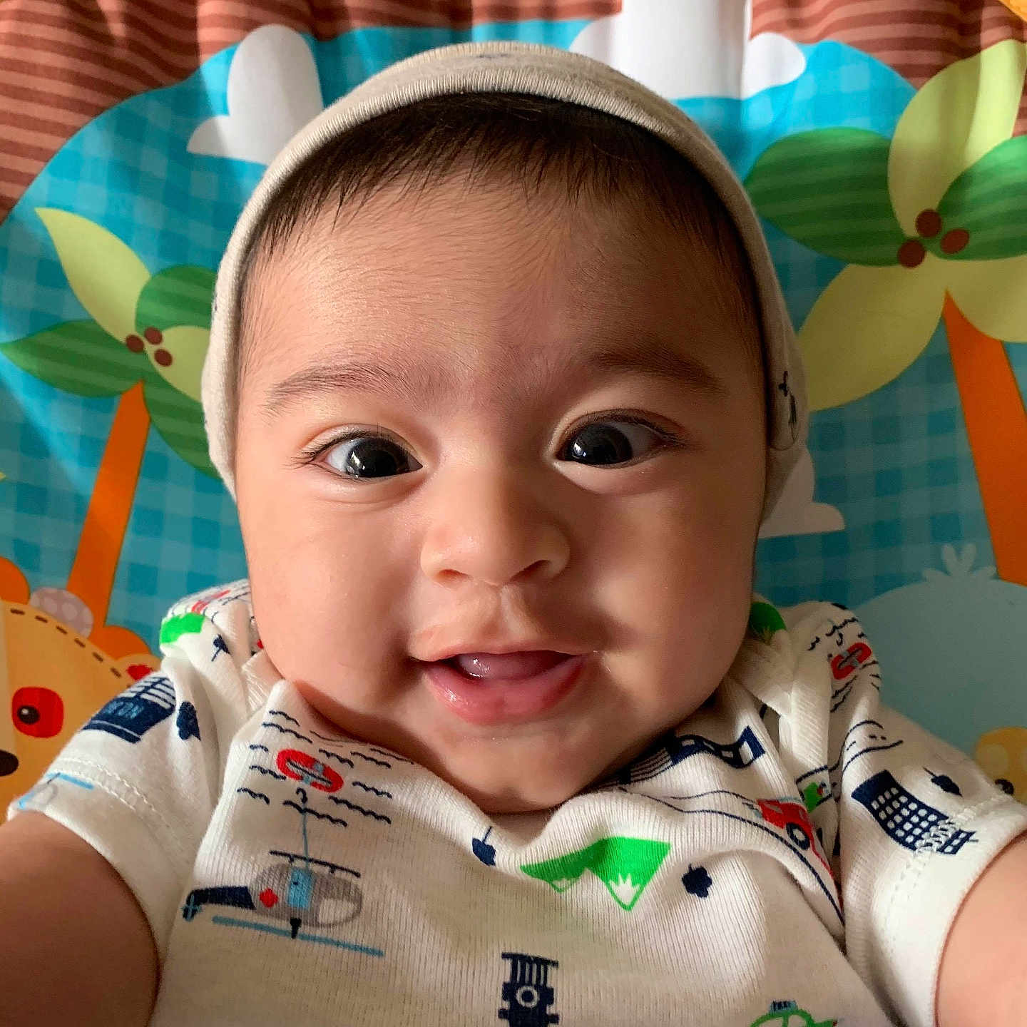 Liam joined the competition — help win amazing prizes! baby, child, smiling, face, hat, onesie, colorful, vehicle_prints, closeup, portrait, infant, happy, cute, indoors, playmat, background, cartoon_flowers, cartoon_animals, young_child, selfie