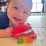 Evan joined the competition — help win amazing prizes! baby, smiling, face, toy, plush_toy, red, colorful, play_mat, indoor, child, happy, cute, hands, closeup, infant, person, bright, natural_light, background, texture