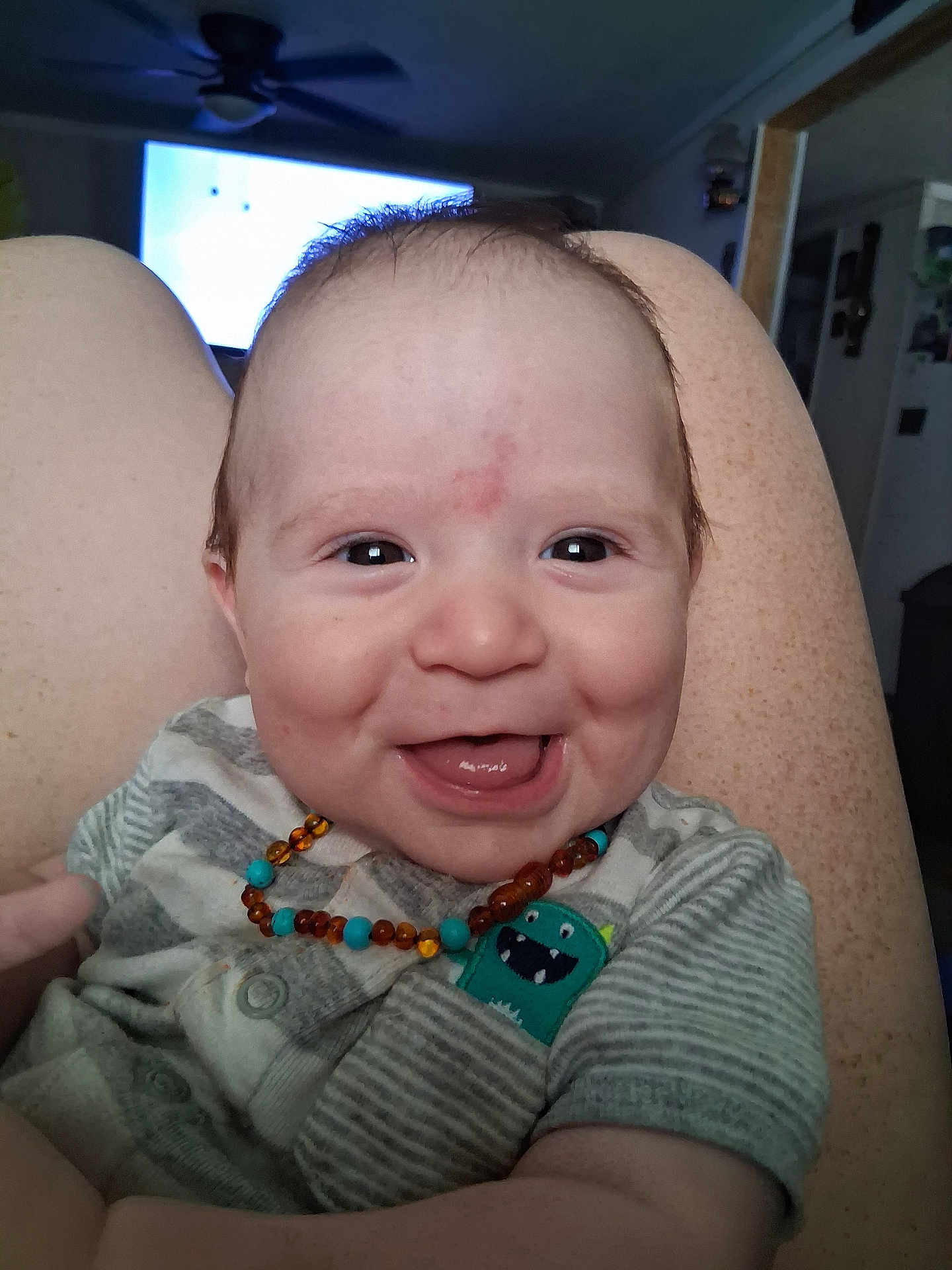 Ledger joined the competition — help win amazing prizes! baby, infant, smiling, laughing, amber_necklace, onesie, striped_clothing, monster_patch, close_up, face, eyes, tongue, cheeks, knees, legs, indoor, living_room, ceiling_fan, television, happy